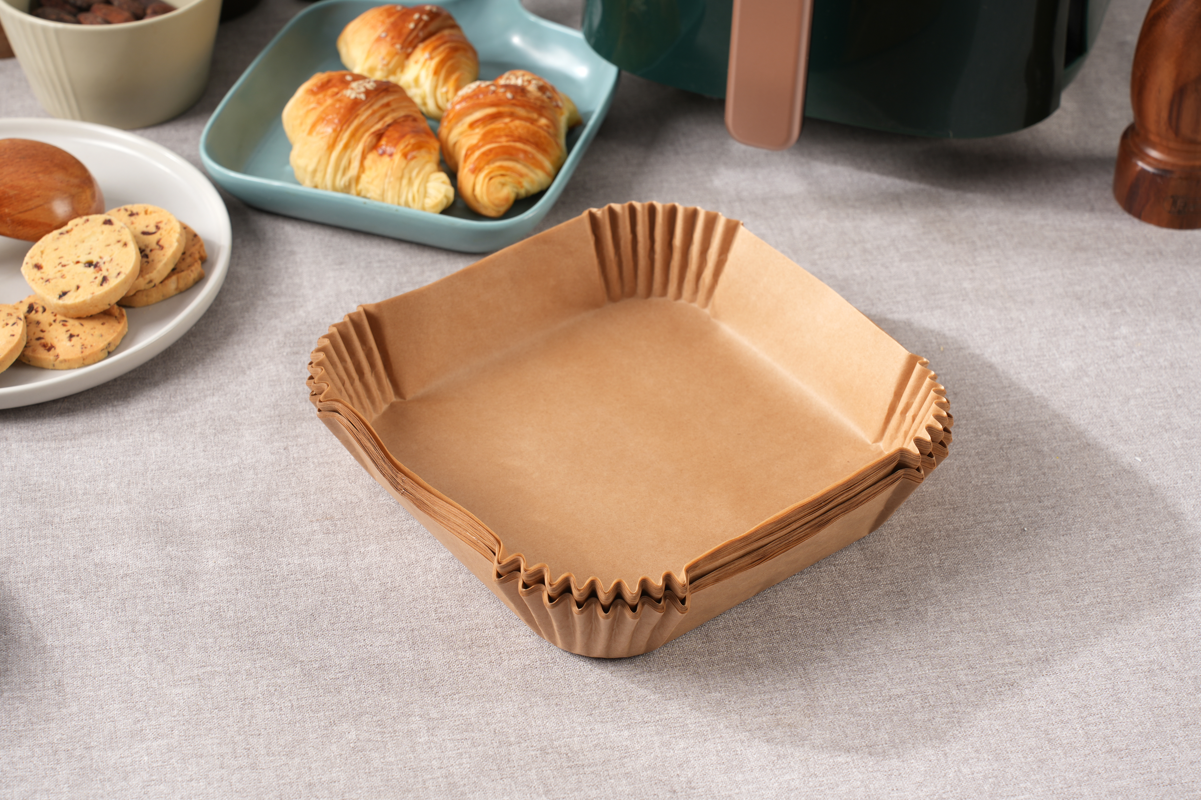 Braune Papierbackformen gestapelt. Kekse und Croissants befinden sich im Hintergrund auf einer grauen Tischdecke.