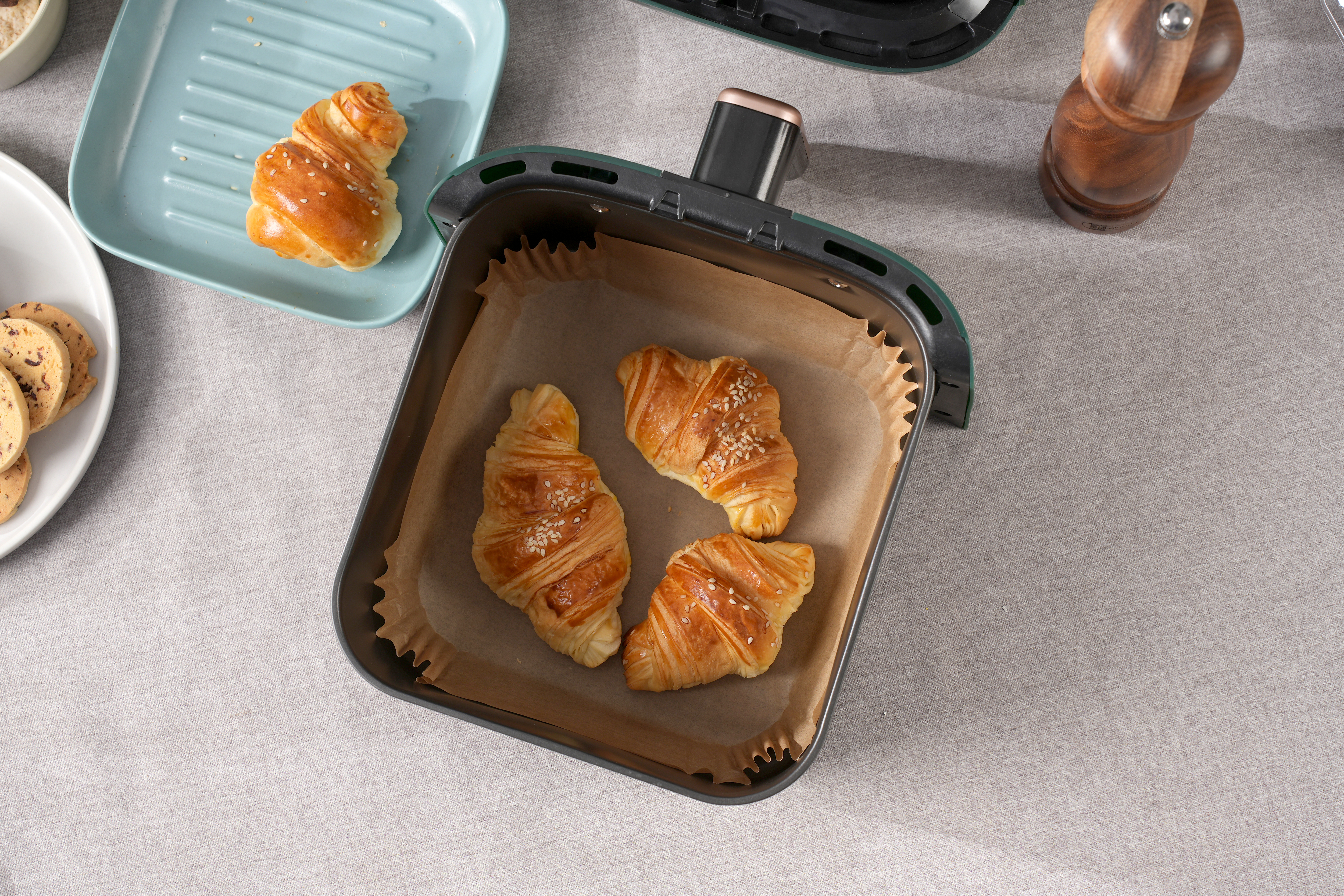 Croissants im Airfryer, einer auf blauem Teller und Kekse links. Graue Tischdecke im Hintergrund.