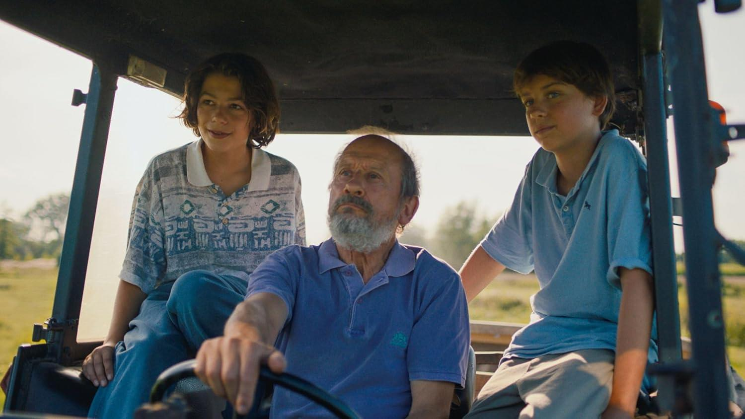 Ein älterer Mann fährt einen Traktor, zwei Jungen sitzen neben ihm auf einem Feld.