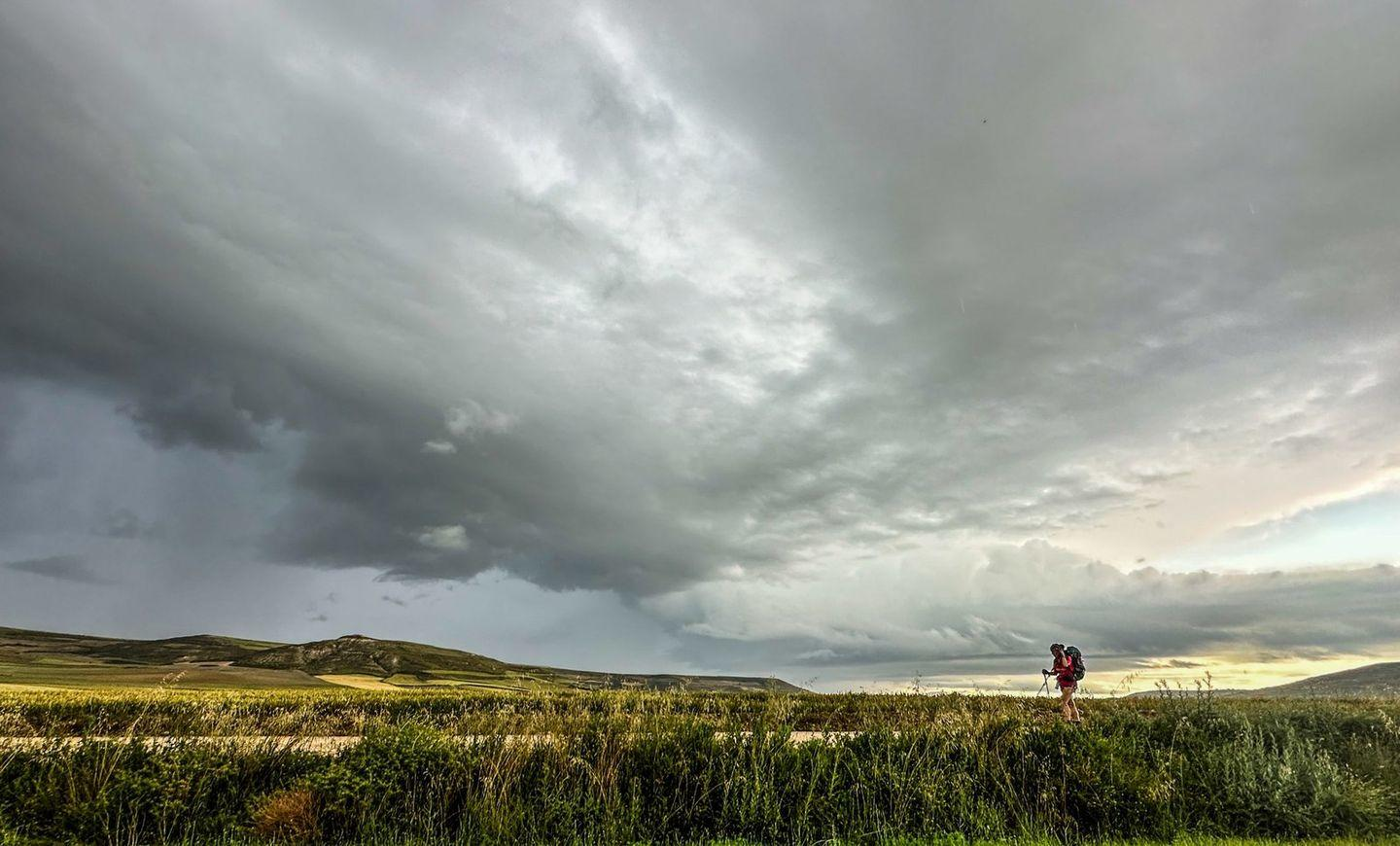 Person geht auf einem Feld unter dunklem, bewölktem Himmel, mit einem Sturm im Hintergrund.