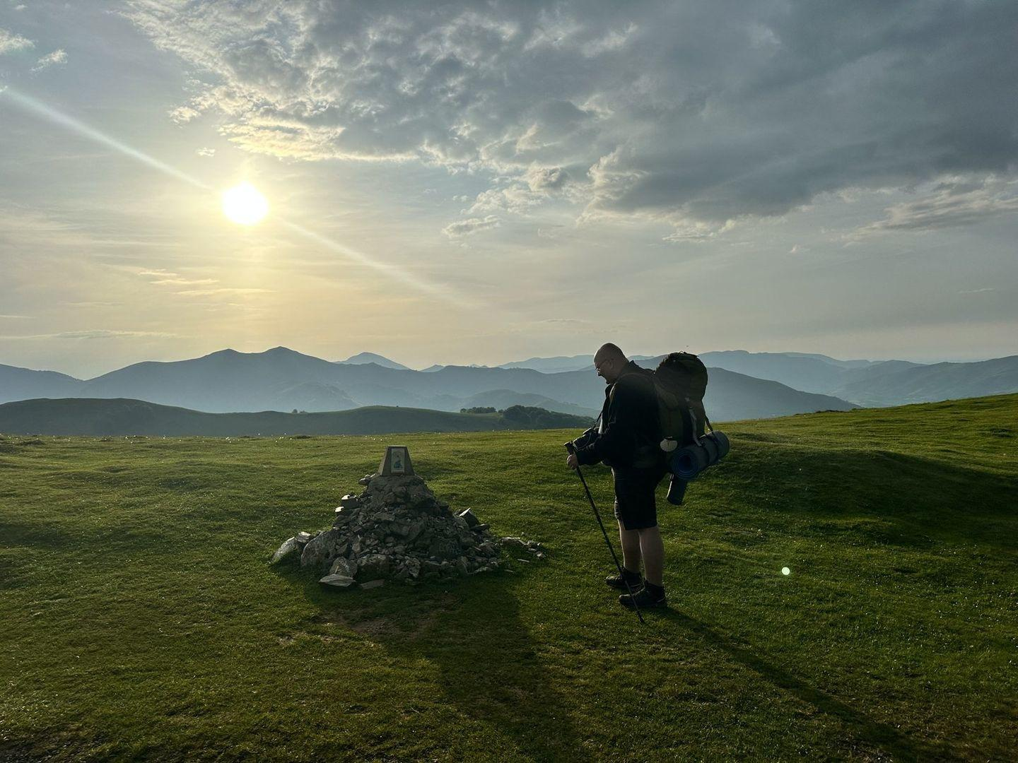 Mann mit Rucksack und Wanderstock steht auf grünem Hügel. Sonnenuntergang, Berge im Hintergrund.