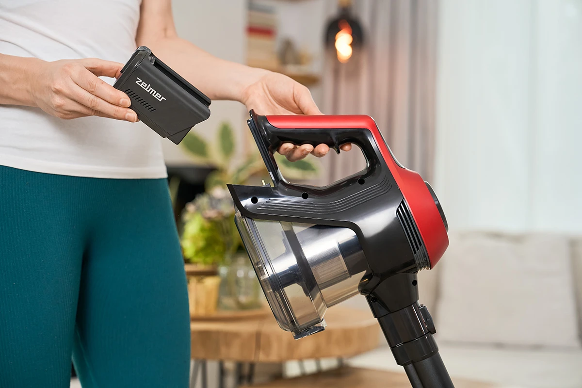 Kobieta wkłada czarną baterię do czerwono-szarego korpusu odkurzacza, w pomieszczeniu.