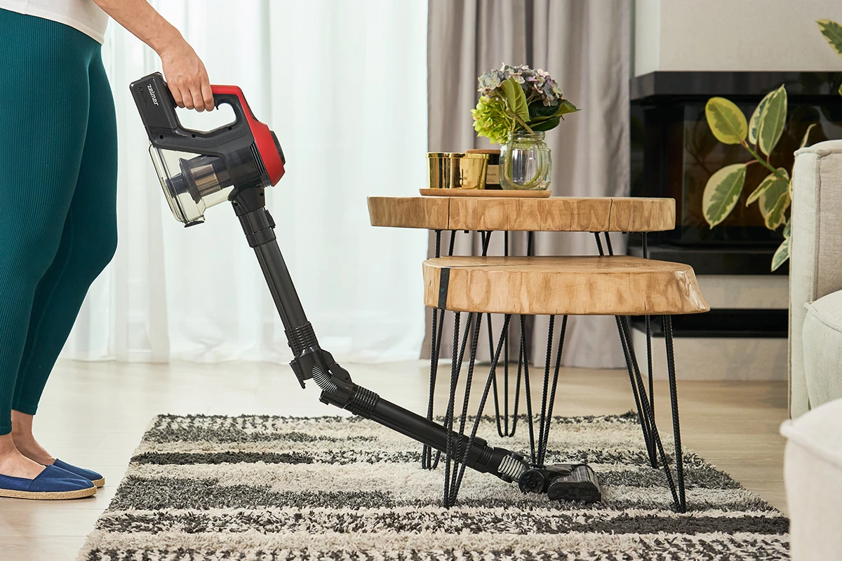 Kobieta w legginsach odkurza dywan czarno-czerwonym odkurzaczem, w pomieszczeniu, z roślinami.