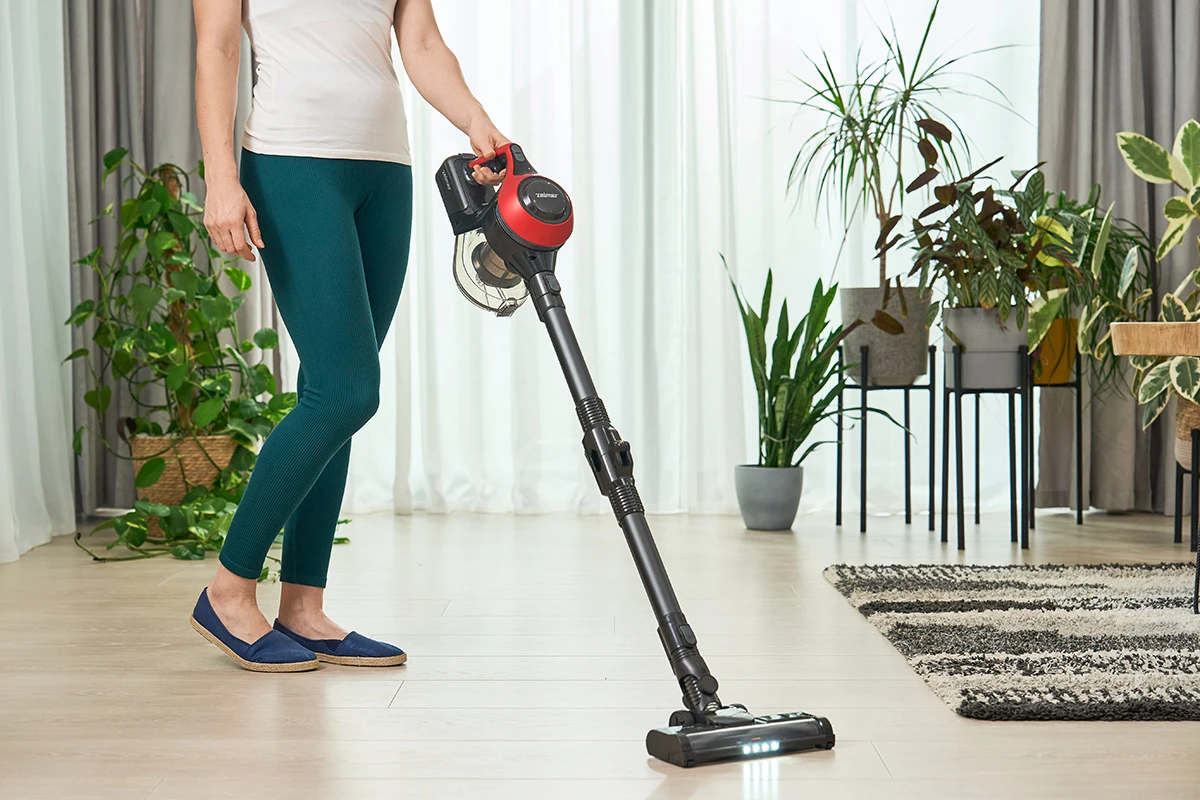 Kobieta odkurza drewnianą podłogę czarno-czerwonym odkurzaczem, w niebieskich butach i legginsach, w pomieszczeniu.