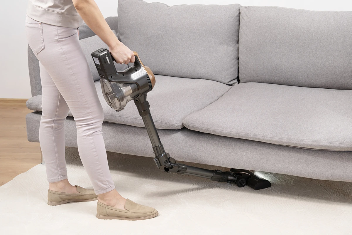 Osoba odkurza pod szarą kanapą odkurzaczem ręcznym na białym dywanie.
