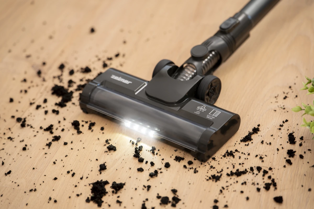 Zbliżenie głowicy odkurzacza z oświetleniem LED na drewnianej podłodze z rozrzuconym brudem.