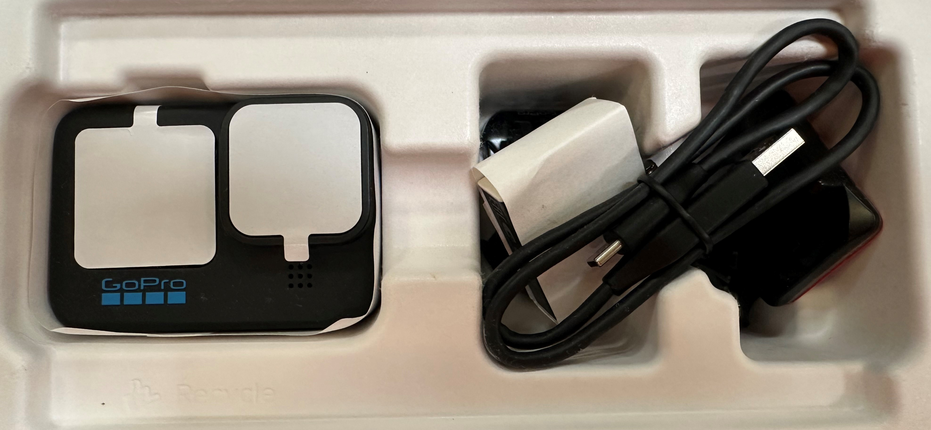 Kamera GoPro i akcesoria w białej tacy. Czarna kamera, kabel do ładowania i biały adapter są widoczne.