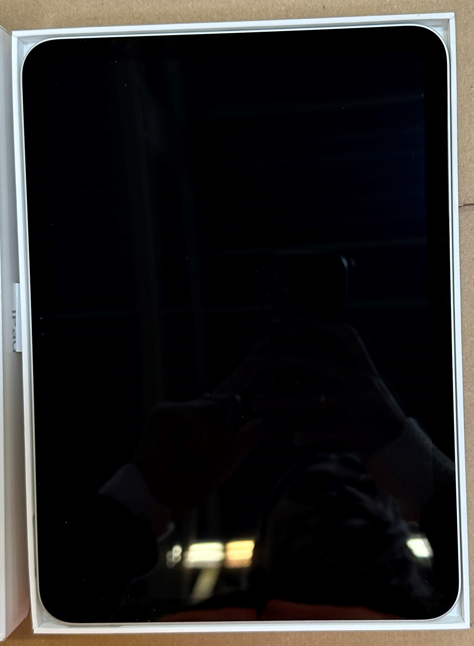 Czarny tablet w białym pudełku, ekranem do góry. Powierzchnia odbijająca, pokazująca odbicie osoby.