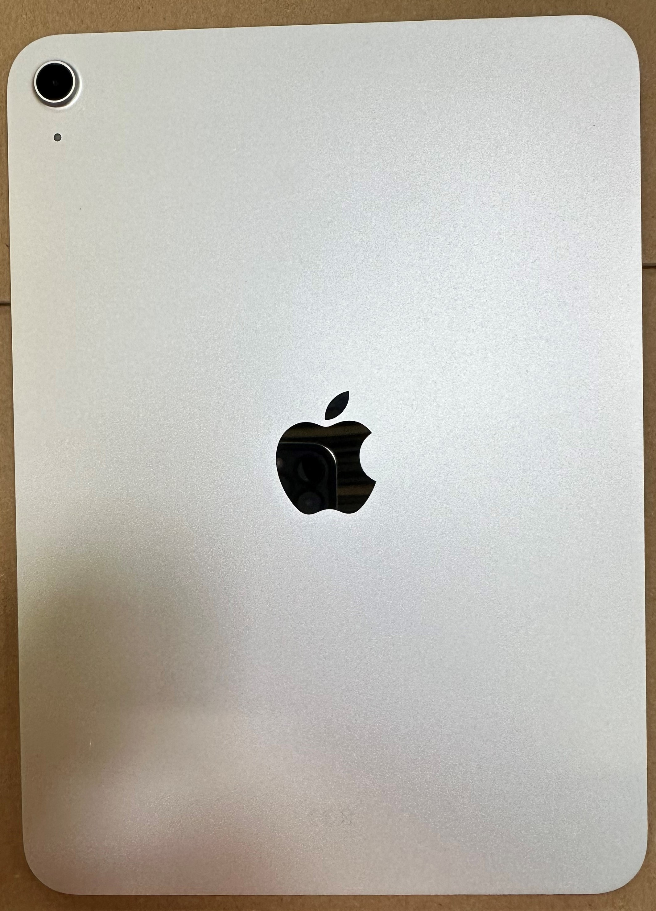 Tył srebrnego tabletu z czarnym logo Apple, aparatem i małą kropką. Brązowa powierzchnia.