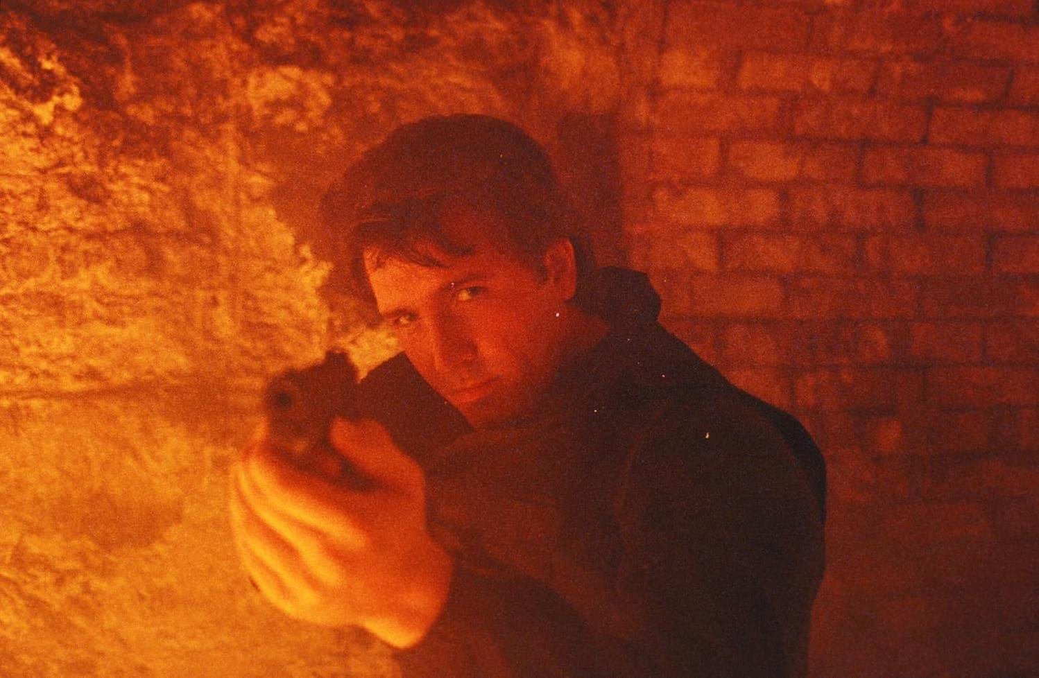 Ein Mann zielt mit einer Waffe in einem dunklen Raum mit orangefarbener Beleuchtung. Er trägt eine schwarze Jacke und schaut in die Kamera.