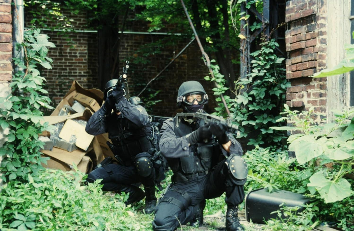 Zwei Personen in schwarzer taktischer Ausrüstung zielen mit Waffen in einem Waldgebiet nahe einem Backsteingebäude.