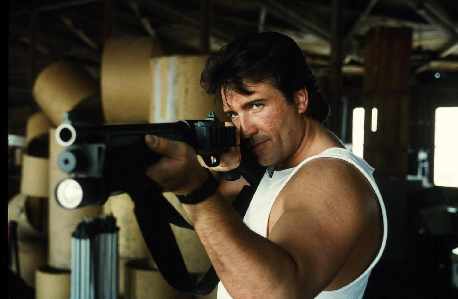 Ein Mann mit einer Schrotflinte, der auf die Kamera zielt. Er trägt ein weißes Tanktop und seine Armmuskeln sind sichtbar.