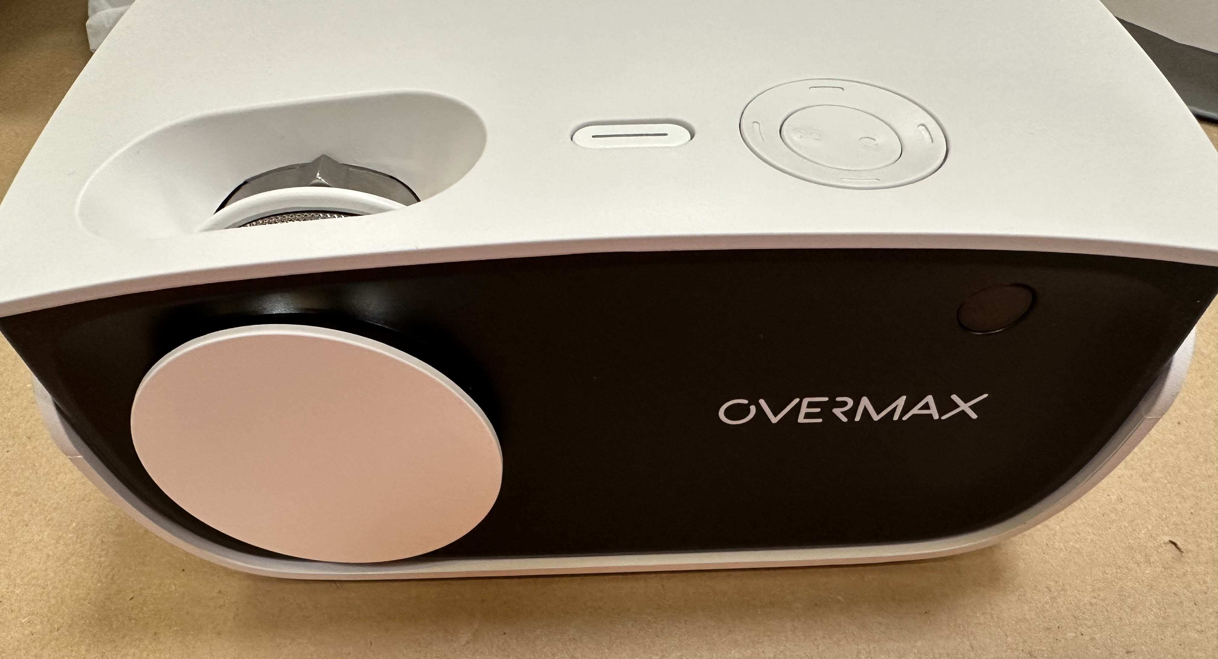 Biało-czarny projektor z napisem OVERMAX. Leży na brązowej powierzchni.