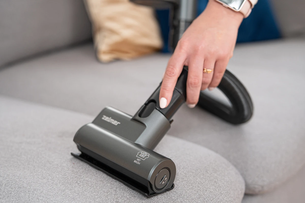 Osoba odkurza szarą sofę za pomocą przystawki do odkurzacza.