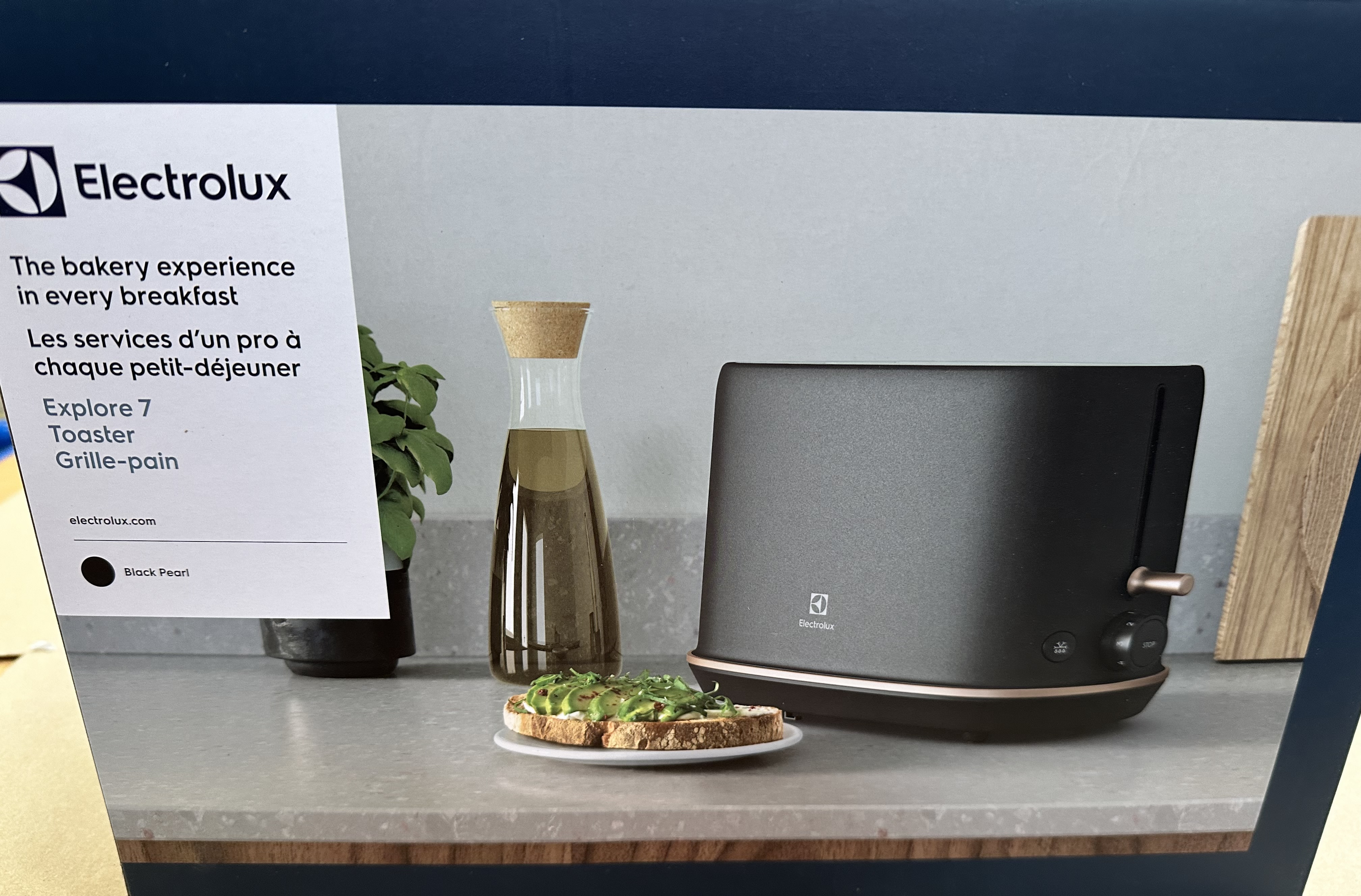 Czarny toster Electrolux z tostami, butelką oleju i rośliną na blacie. Na pudełku napisano "Doświadczenie piekarni w każdym śniadaniu".