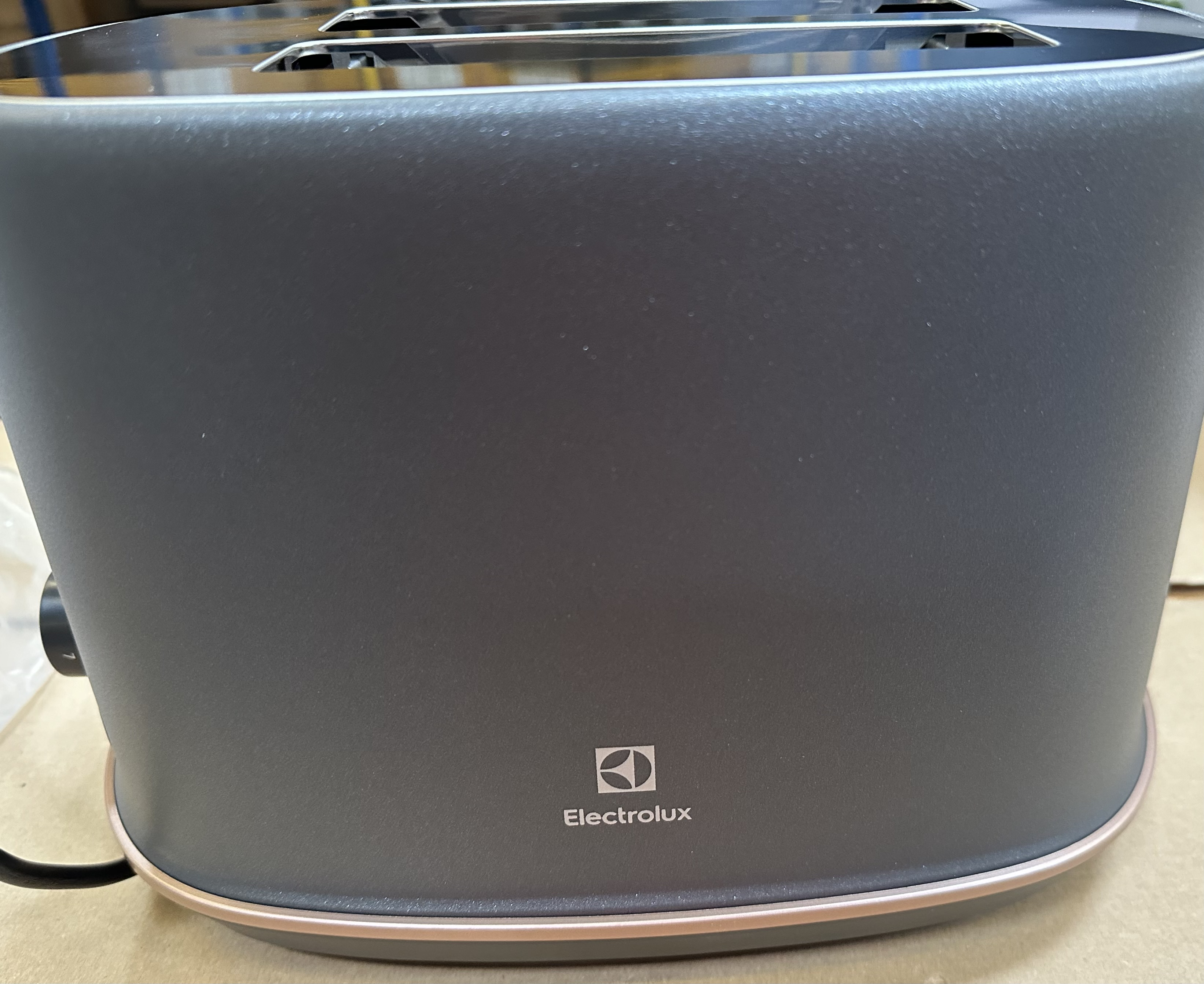 Zbliżenie na czarny toster Electrolux z podstawą w kolorze różowego złota. Logo jest widoczne. Toster stoi na jasnobrązowej powierzchni.