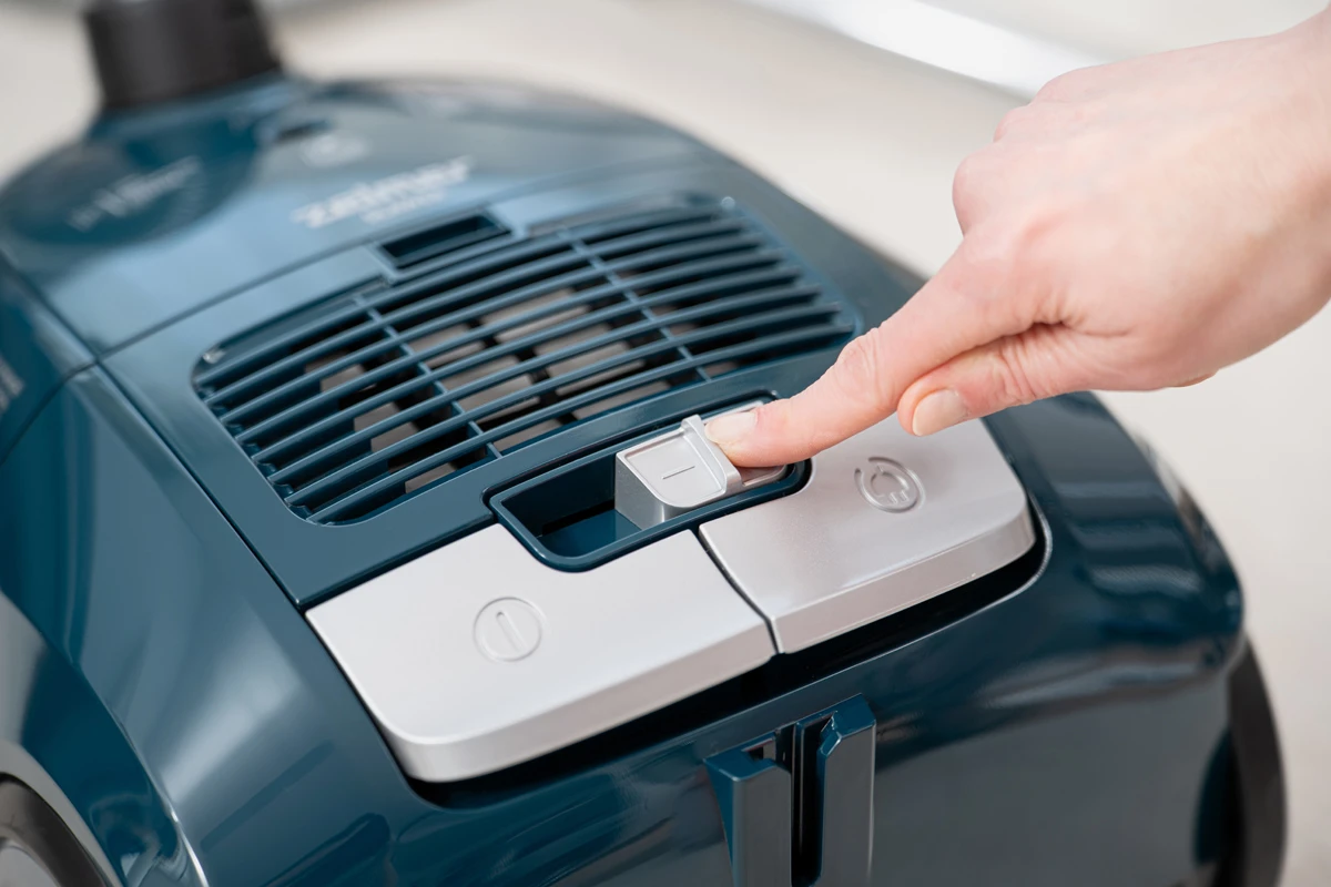 Dłoń naciska przycisk na turkusowym odkurzaczu, zbliżenie, pokazujące szczegóły przełącznika.