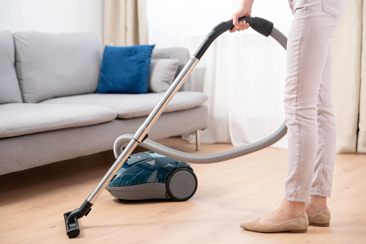 Osoba odkurza drewnianą podłogę turkusowym odkurzaczem, obok szarej kanapy.
