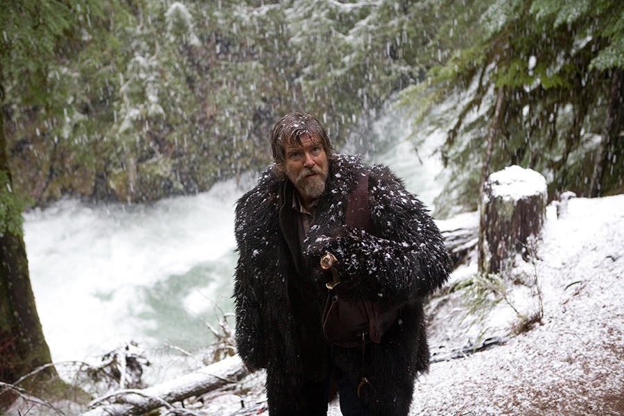 Ein Mann steht in einem verschneiten Wald und trägt einen Pelzmantel, im Hintergrund ein Wasserfall.