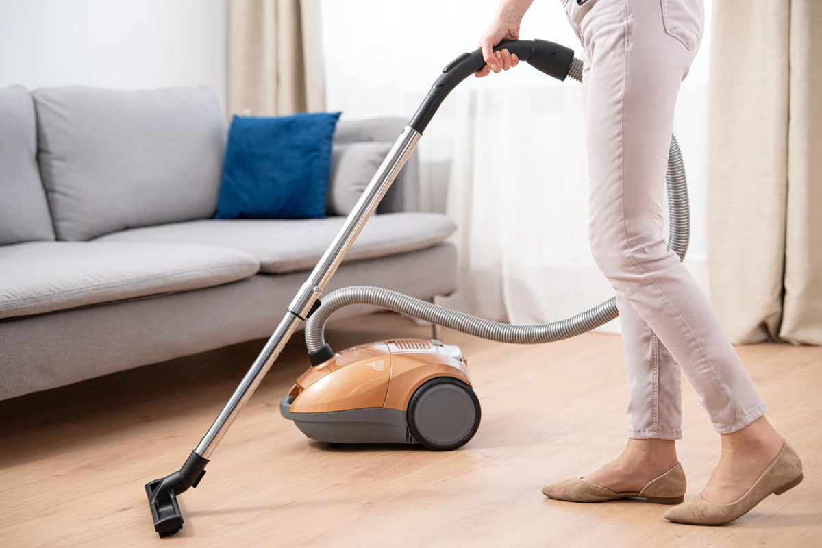 Osoba odkurza drewnianą podłogę. W tle kanapa i niebieska poduszka.