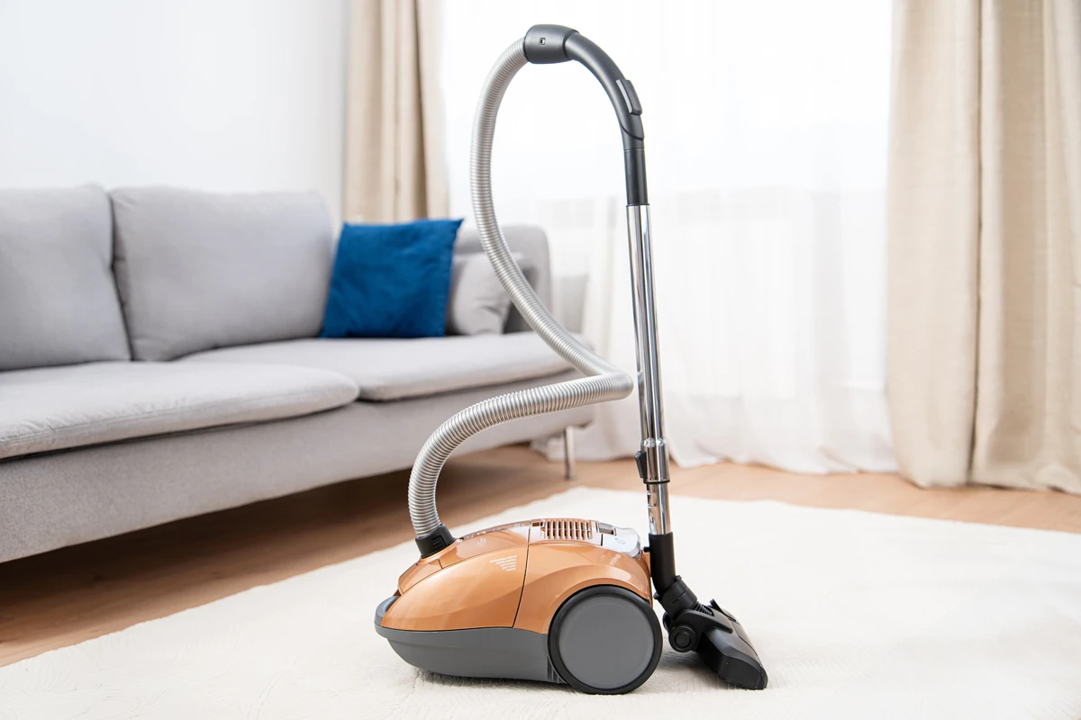 Odkurzacz stoi na białym dywanie. W tle kanapa i niebieska poduszka.