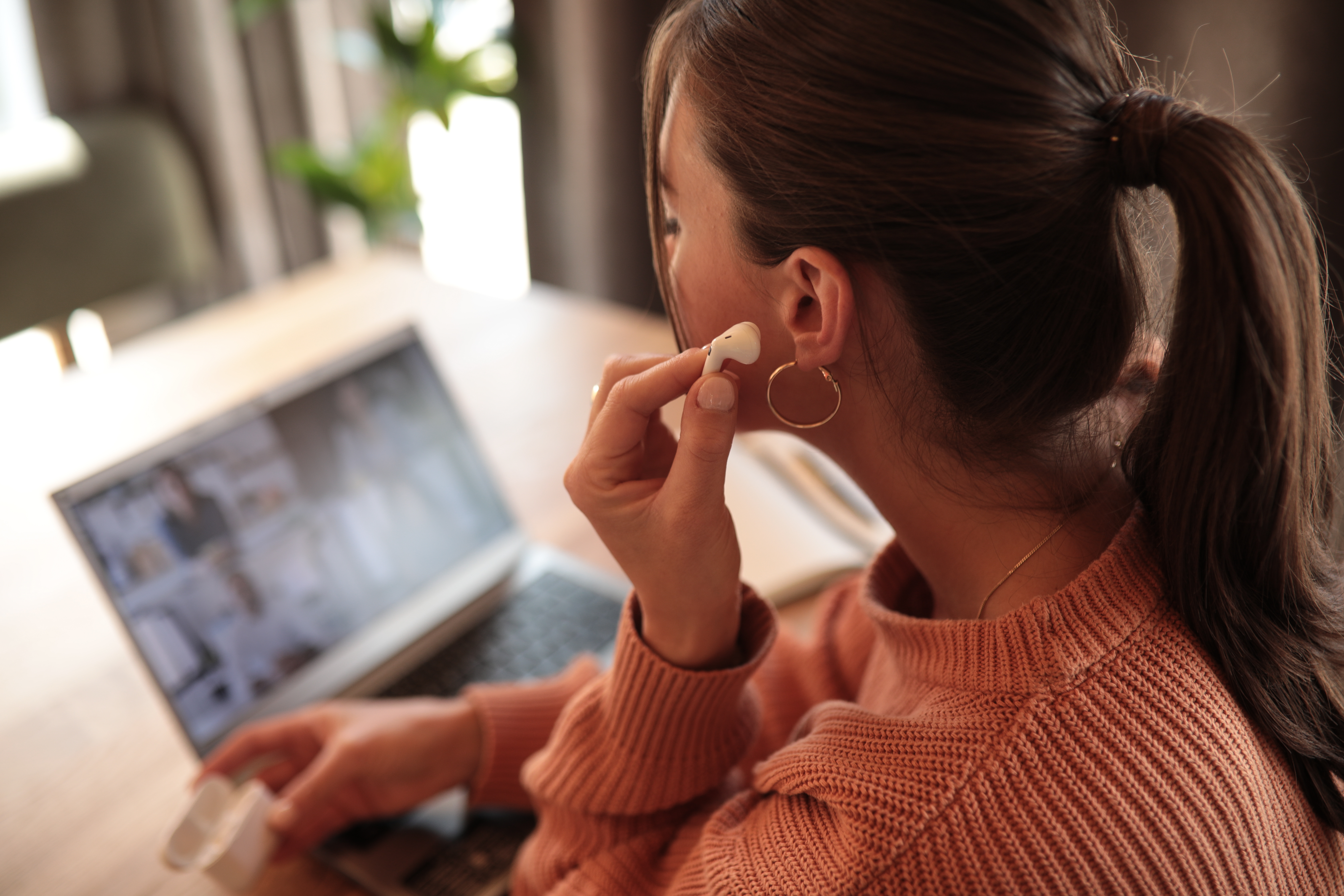 Eine Frau in einem rosa Pullover, die einen Ohrhörer trägt, schaut auf einen Laptop.
