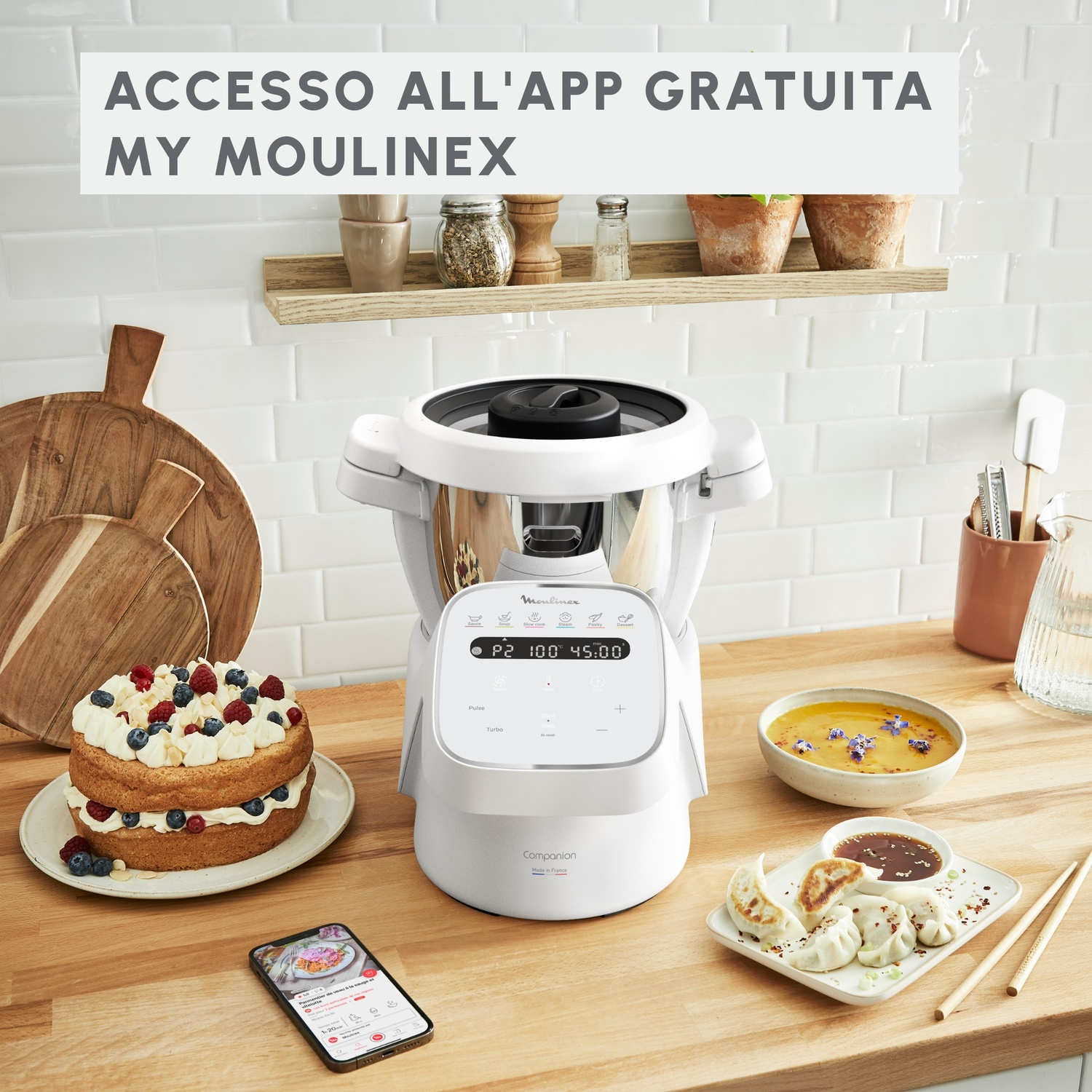 Robot da cucina Moulinex bianco con torta, ravioli e telefono su un tavolo di legno.