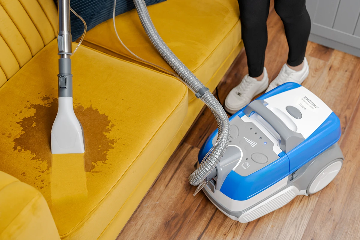 Zabytkowa pralka czyszcząca żółtą sofę, z osobą w pobliżu.