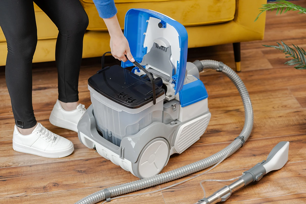 Osoba otwiera niebiesko-szary odkurzacz. Żółta sofa i drewniana podłoga w tle.