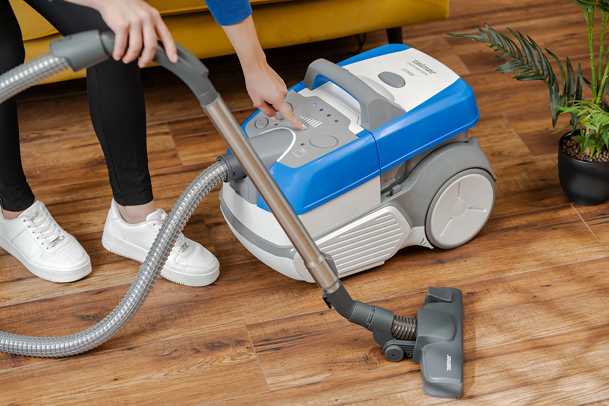 Osoba obsługuje niebiesko-biały odkurzacz na drewnianej podłodze. Odkurzacz znajduje się w pobliżu małej rośliny doniczkowej.