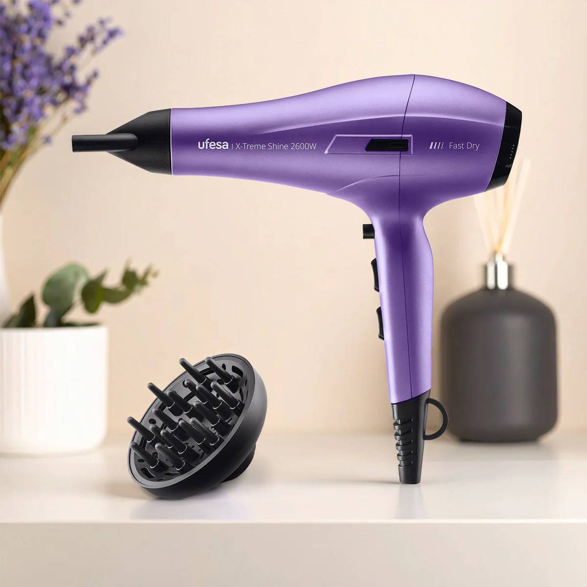 Secador de pelo morado con difusor y accesorios en una mesa blanca. Jarrón y difusor en el fondo.