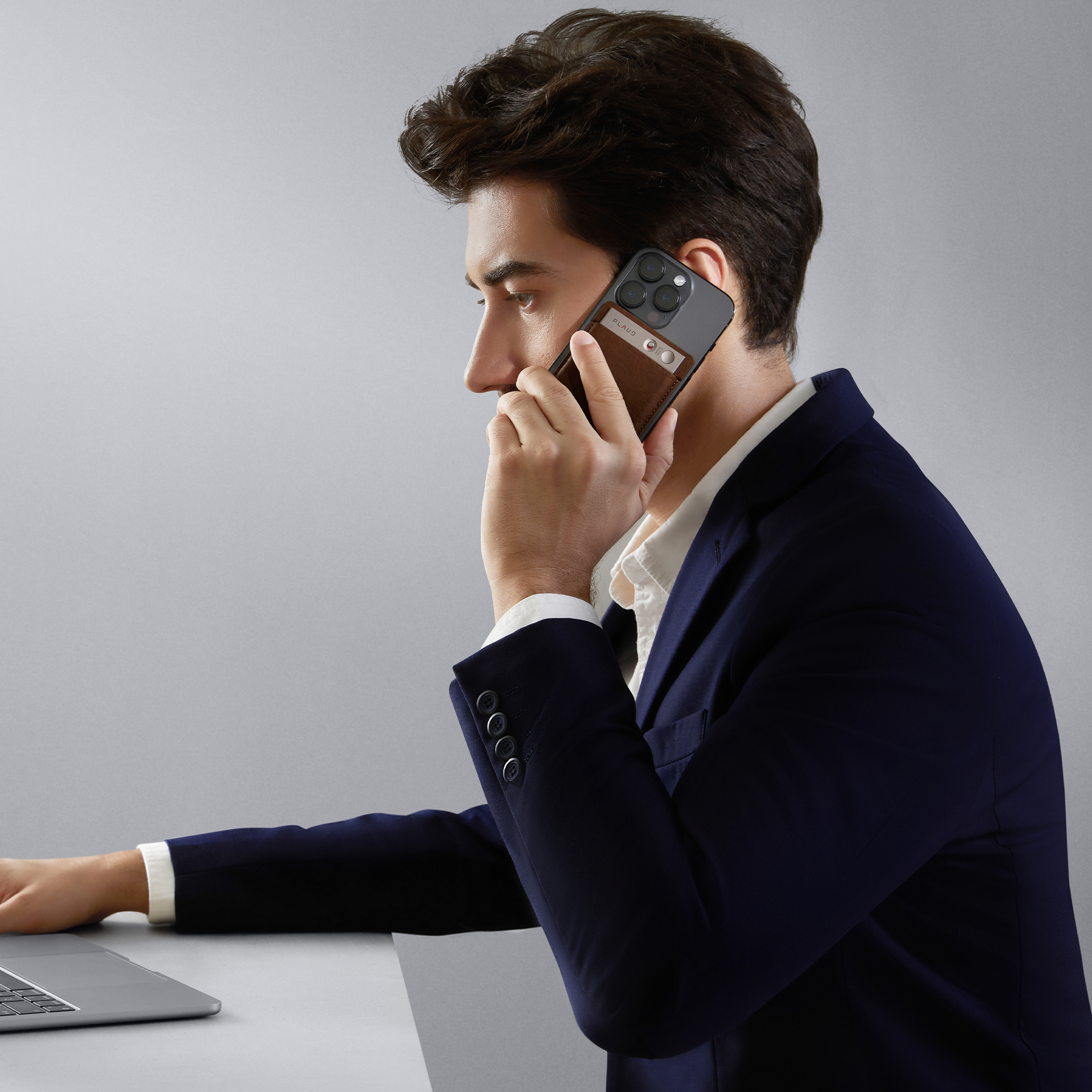 Ein Mann hält ein Telefon mit brauner Lederhülle, spricht und arbeitet am Laptop.