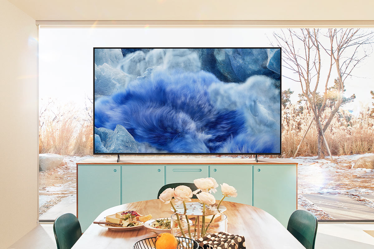 Un televisore mostra un'immagine di nuvola blu, posta sopra un tavolo. Un tavolo con cibo e fiori è davanti.