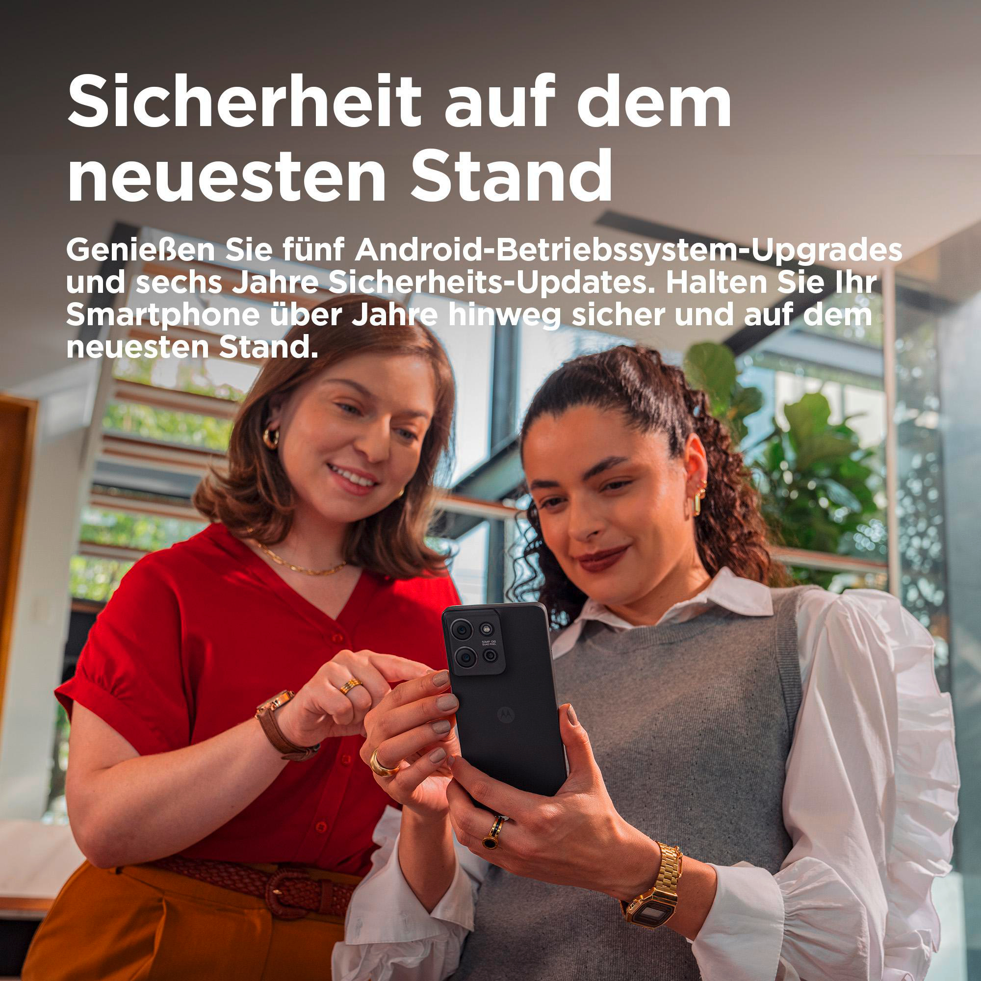 Zwei Frauen schauen auf ein schwarzes Telefon. Eine trägt ein rotes Hemd, die andere einen grauen Pullover.