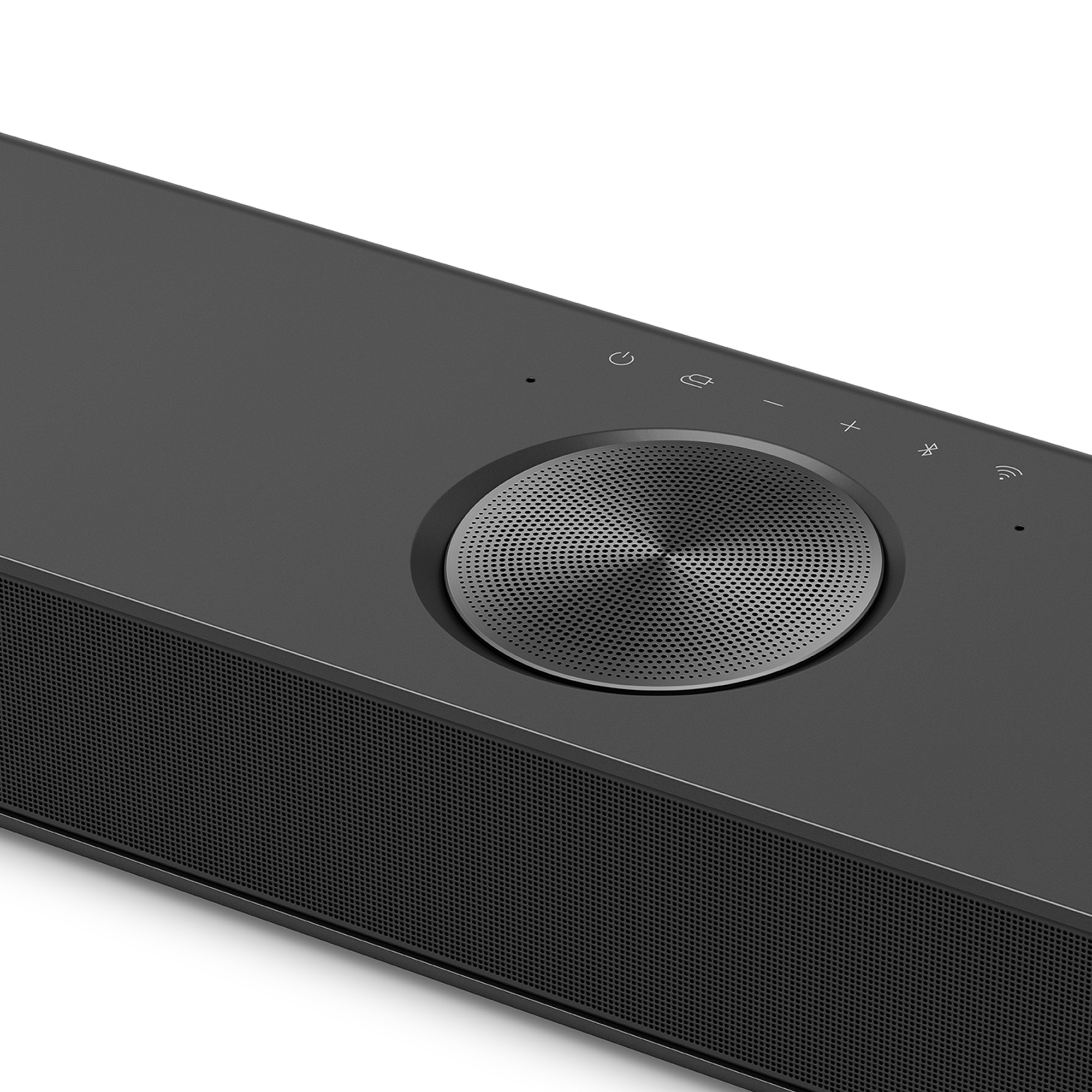 Primer plano de los controles de la barra de sonido. Disco plateado con botones de encendido, volumen y selección de fuente, y una rejilla de altavoz de malla.