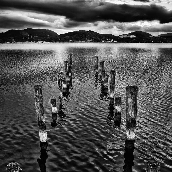 Schwarz-Weiß-Foto von Holzpfählen im Wasser, die zu Bergen unter einem bewölkten Himmel führen.