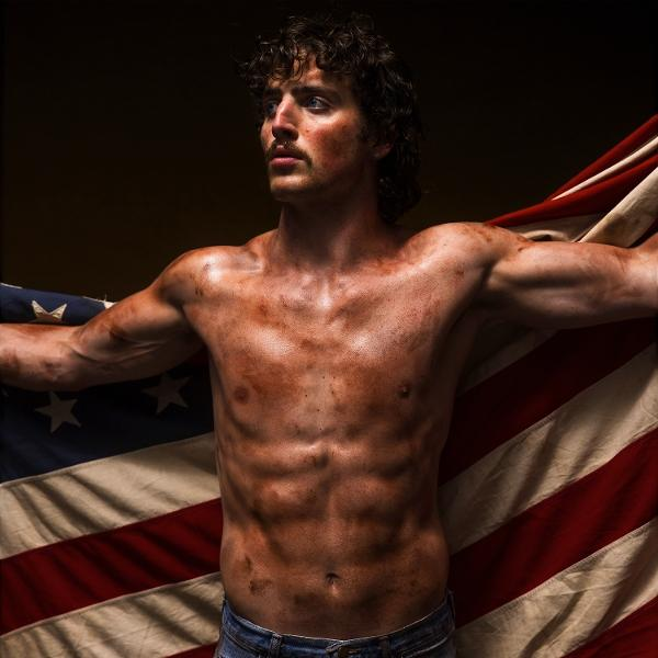 Ein Mann ohne Hemd mit einer amerikanischen Flagge. Er hat einen Schnurrbart und ist mit Schmutz bedeckt.