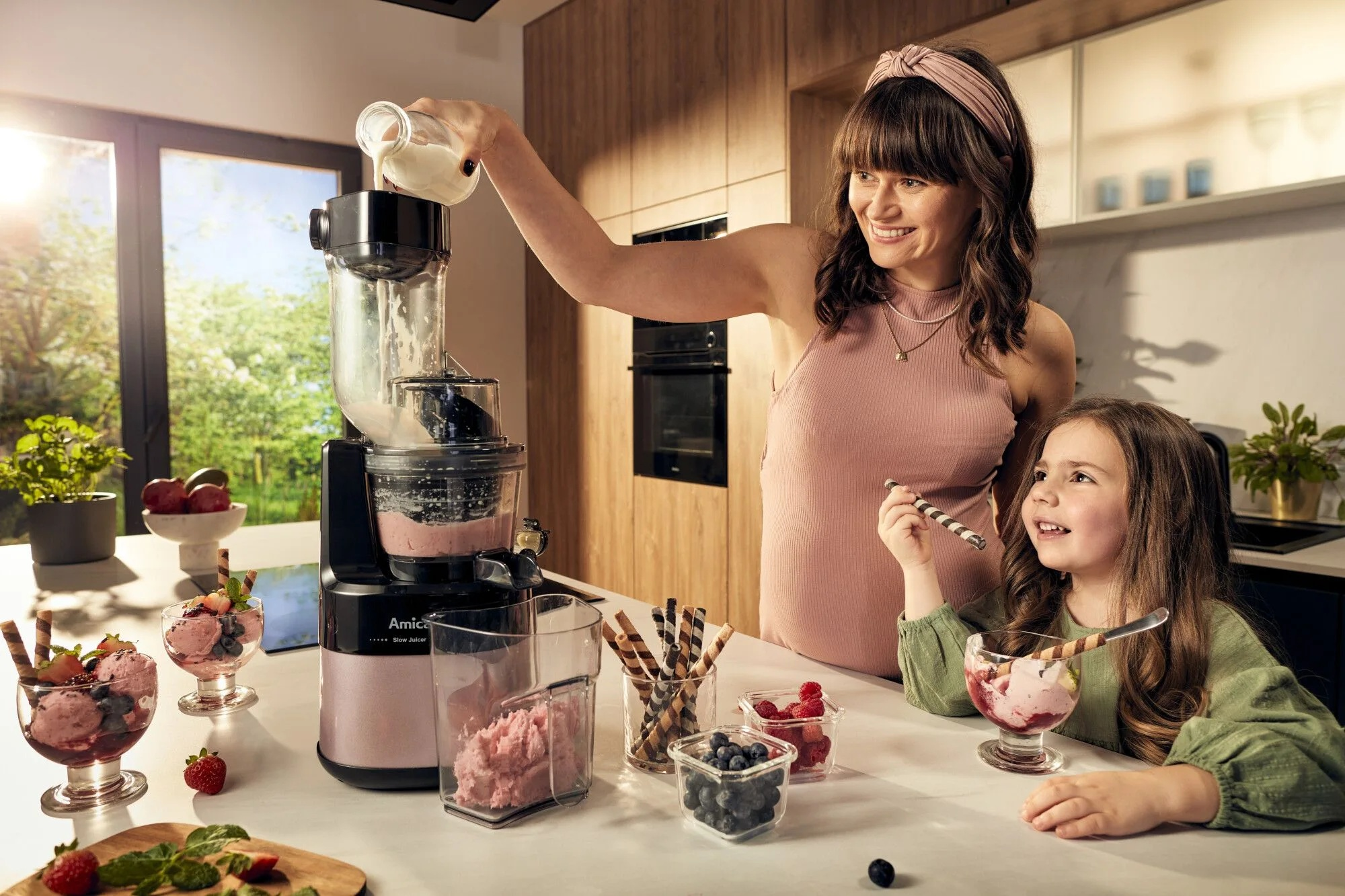 Kobieta wlewa płyn do wyciskarki, z dzieckiem i deserami w tle.