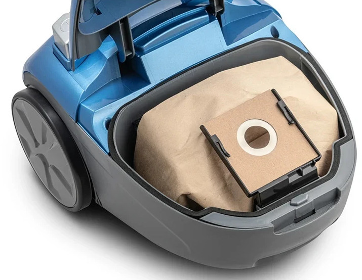 Niebieski odkurzacz z otwartą komorą. W środku papierowy worek z filtrem.