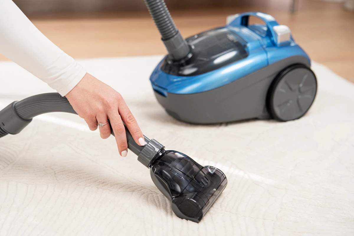 Ręka obsługuje odkurzacz na białym dywanie. Niebieski odkurzacz w tle, drewniana podłoga.