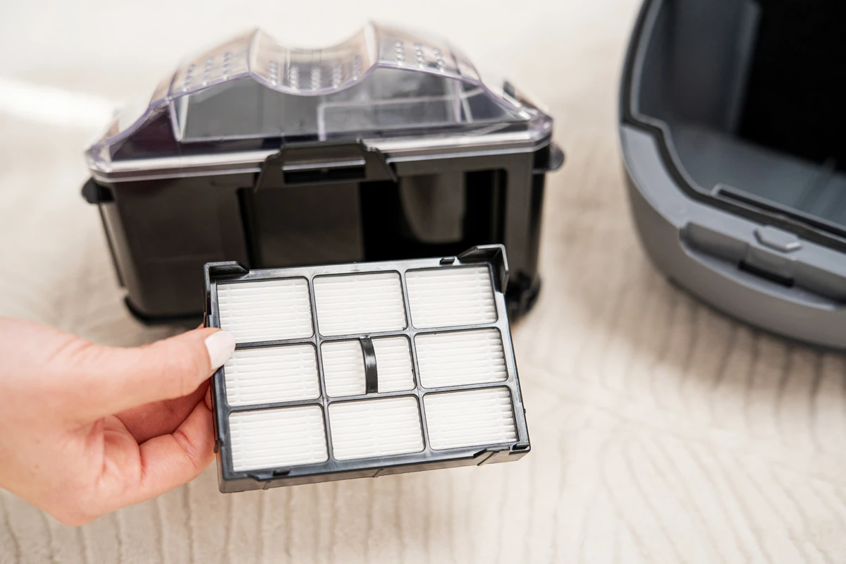 Ręka trzymająca filtr do odkurzacza. Czarne części odkurzacza i szary element w tle na jasnej powierzchni.