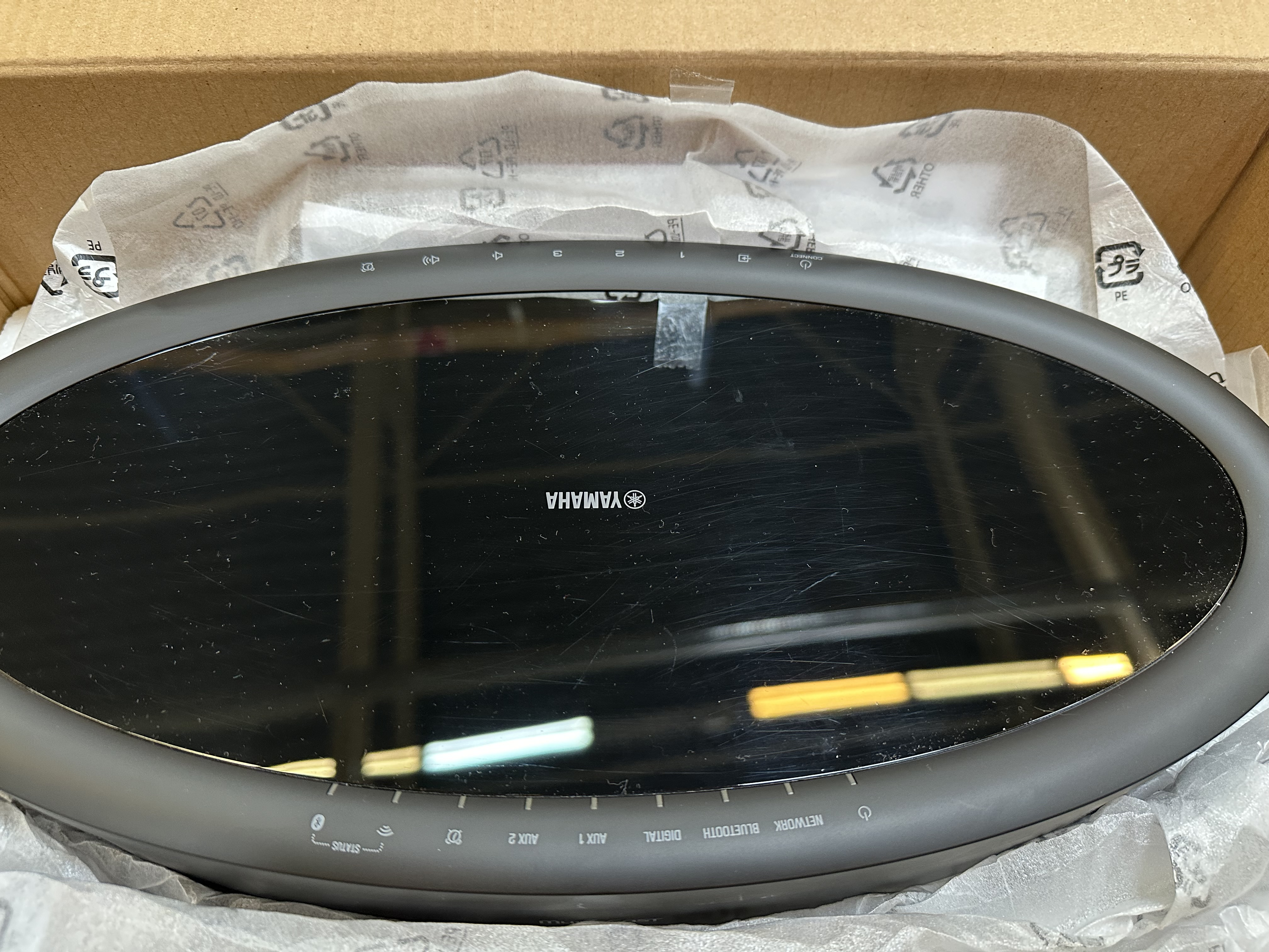 Czarny soundbar Yamaha jest w pudełku. Ma błyszczący ekran i ciemną ramę. Pudełko jest brązowe z białym papierem do pakowania.