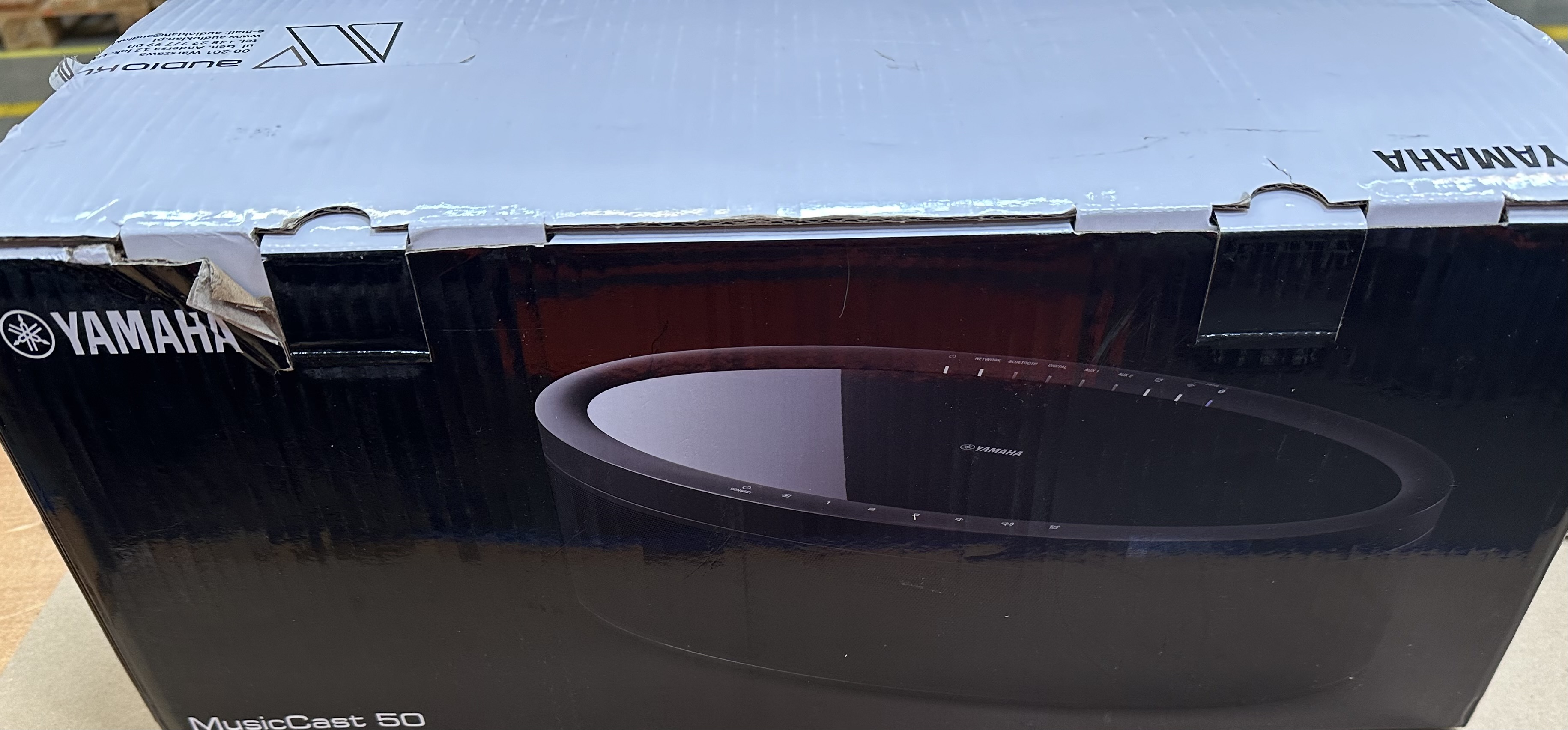 Pokazano pudełko głośnika Yamaha MusicCast 50. Pudełko jest czarne, z okrągłym głośnikiem. Widoczne jest logo Yamaha.