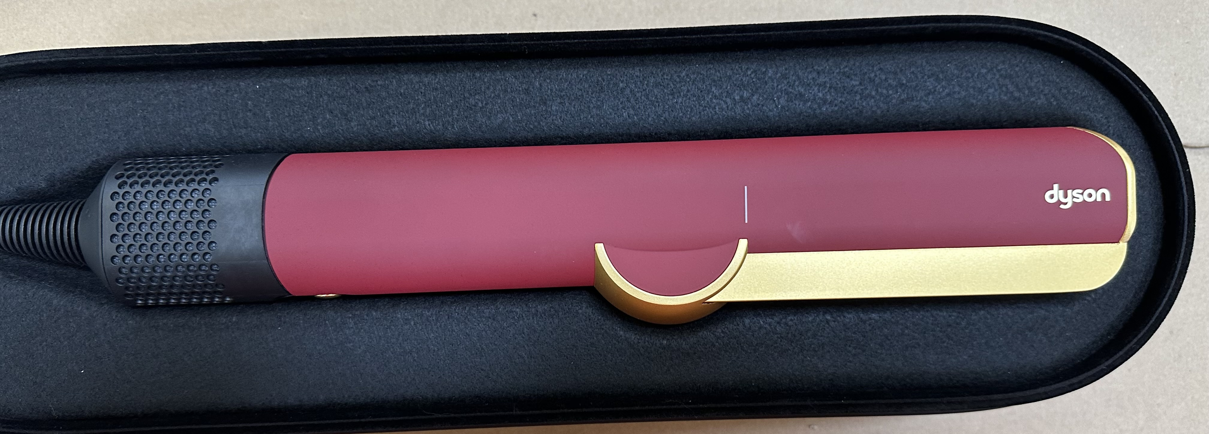 Czerwono-złote narzędzie do włosów Dyson spoczywa w czarnym etui. Narzędzie jest bordowe ze złotymi akcentami.