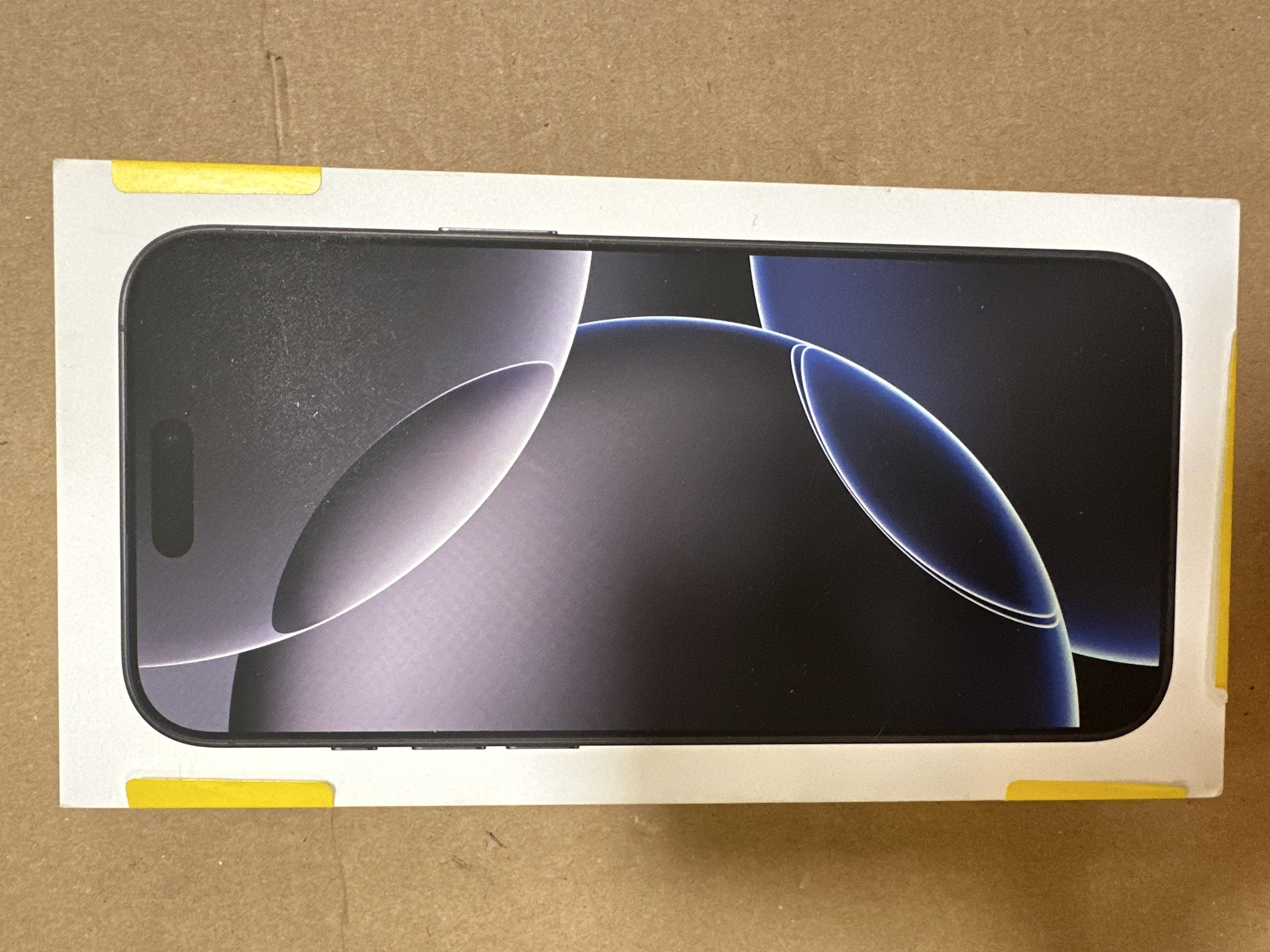 Pudełko iPhone'a na brązowej powierzchni. Pudełko jest białe z obrazem ekranu. Żółta taśma uszczelnia pudełko.