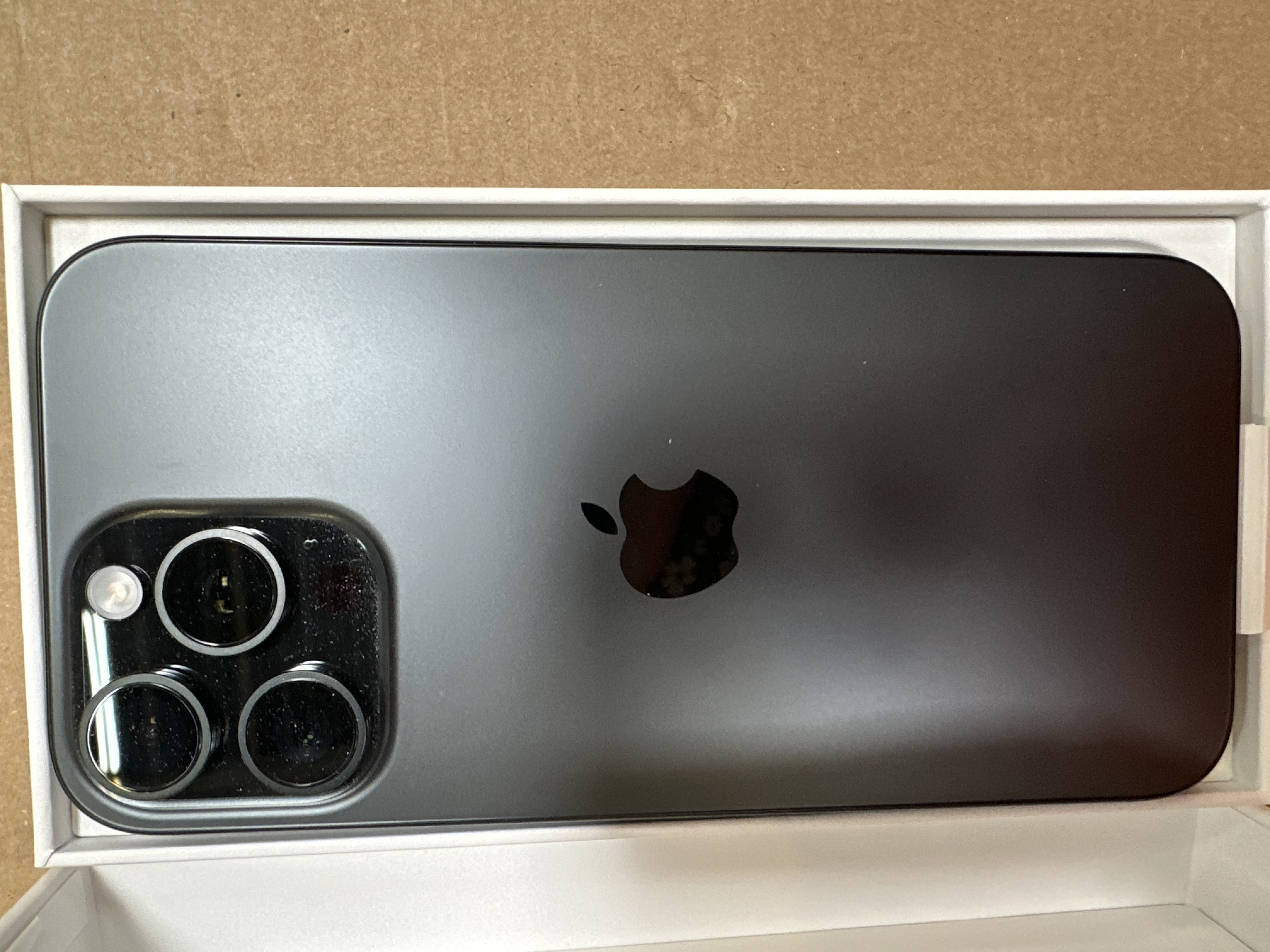 Ciemnoszary iPhone w białym pudełku. Tył pokazuje kamery, logo Apple i powierzchnię telefonu.