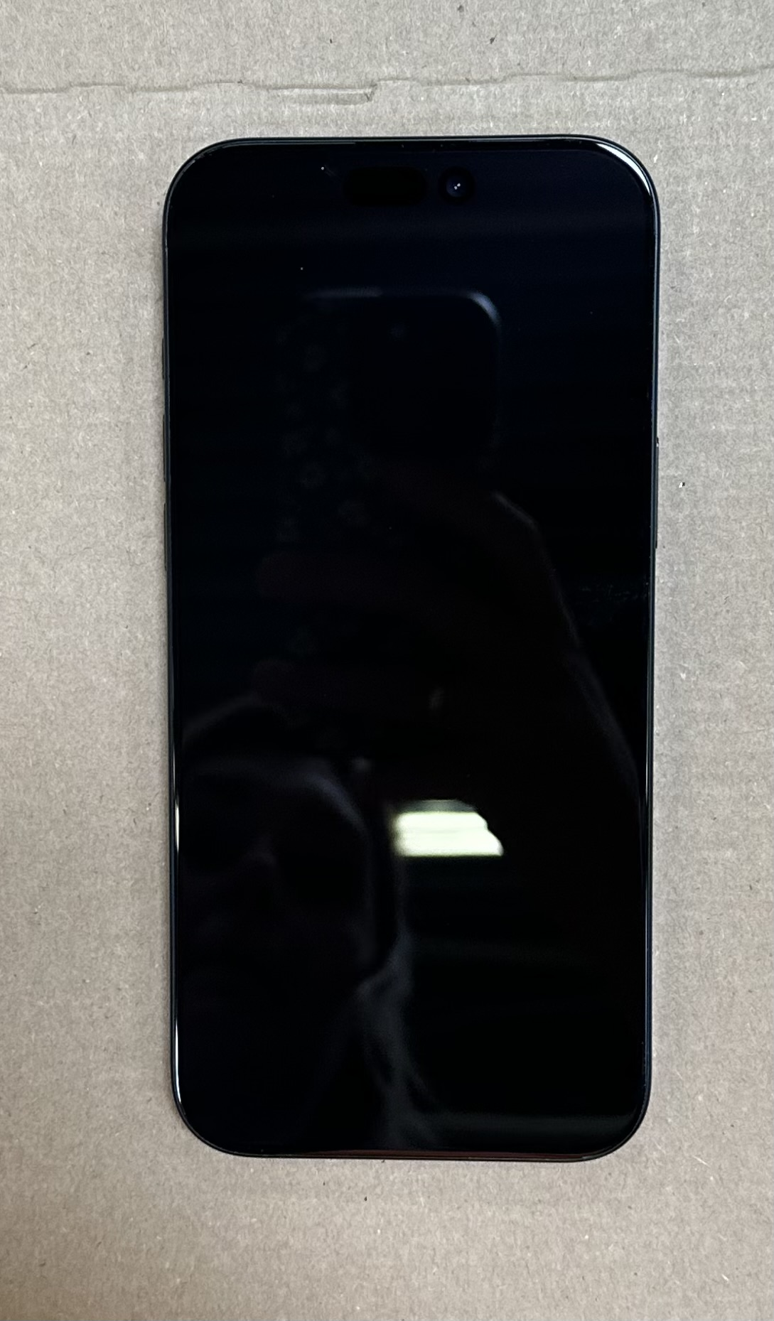 Czarny smartfon z pustym ekranem, odbijającym światło. Leży na szarej tekturze. Widoczny aparat, czujniki u góry.