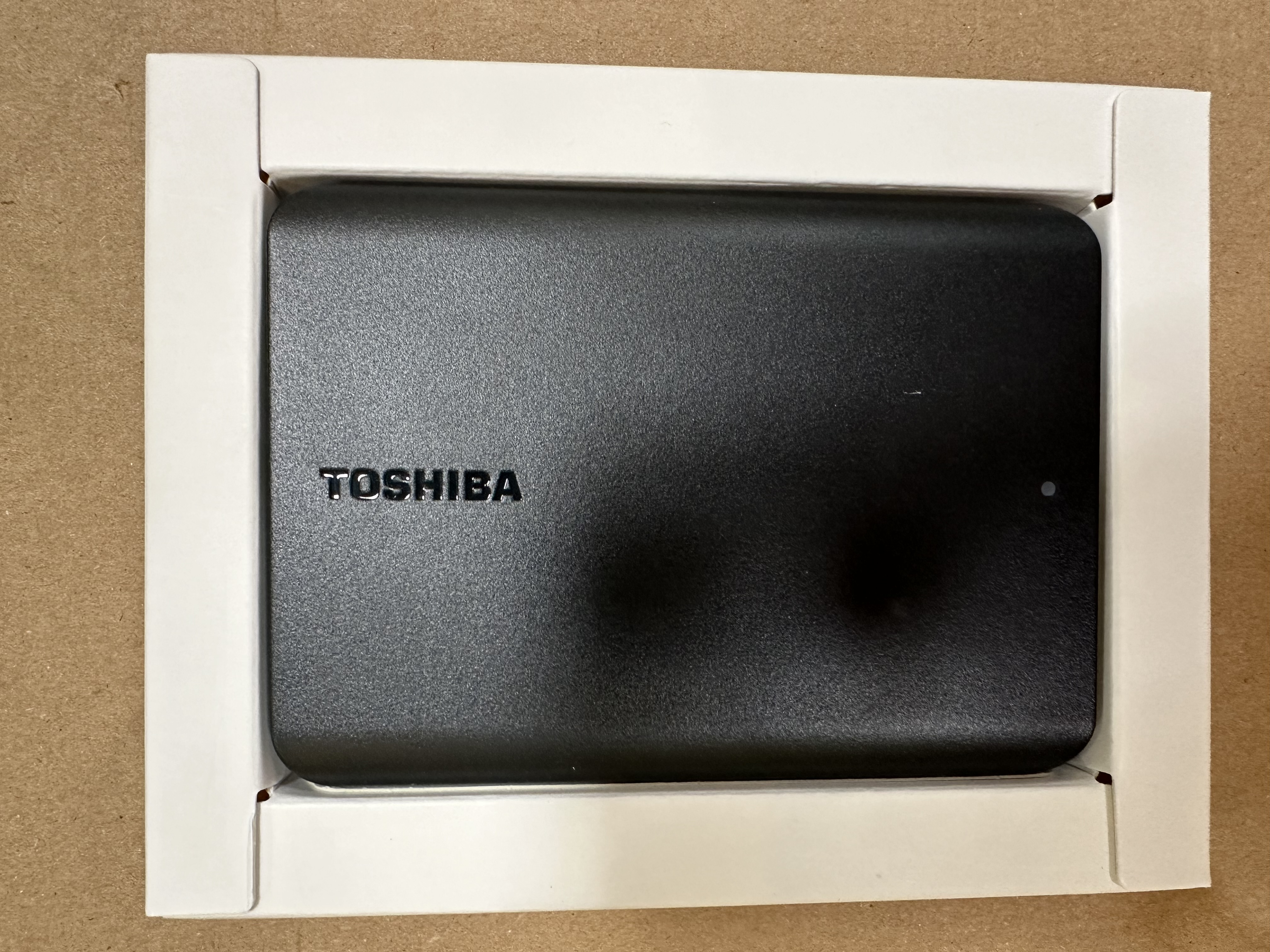 Czarny dysk twardy zewnętrzny Toshiba w białej kartonowej ramce na brązowej powierzchni.