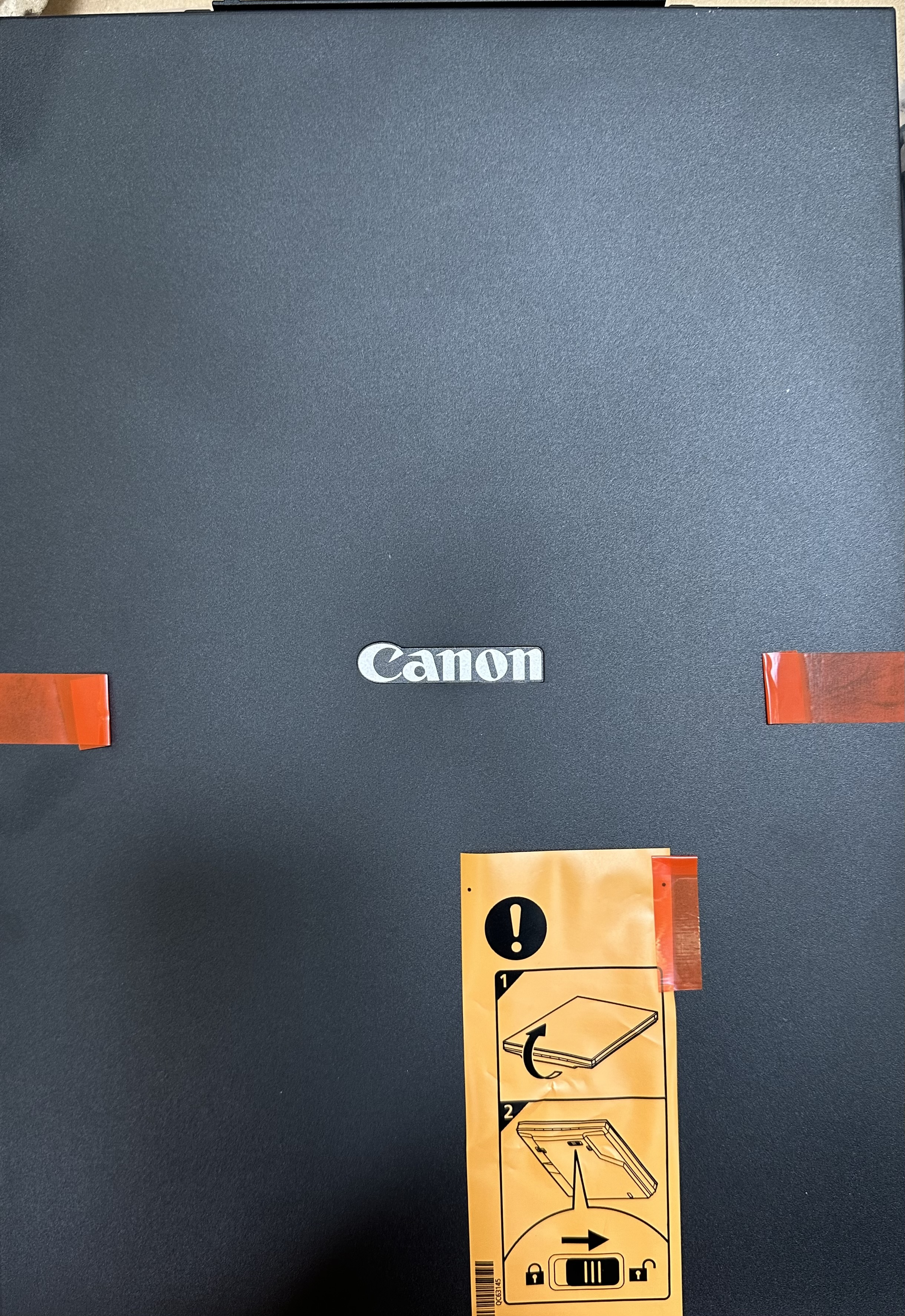 Czarna drukarka z logo Canon. Pomarańczowa taśma na maszynie. Instrukcje na żółtej naklejce.