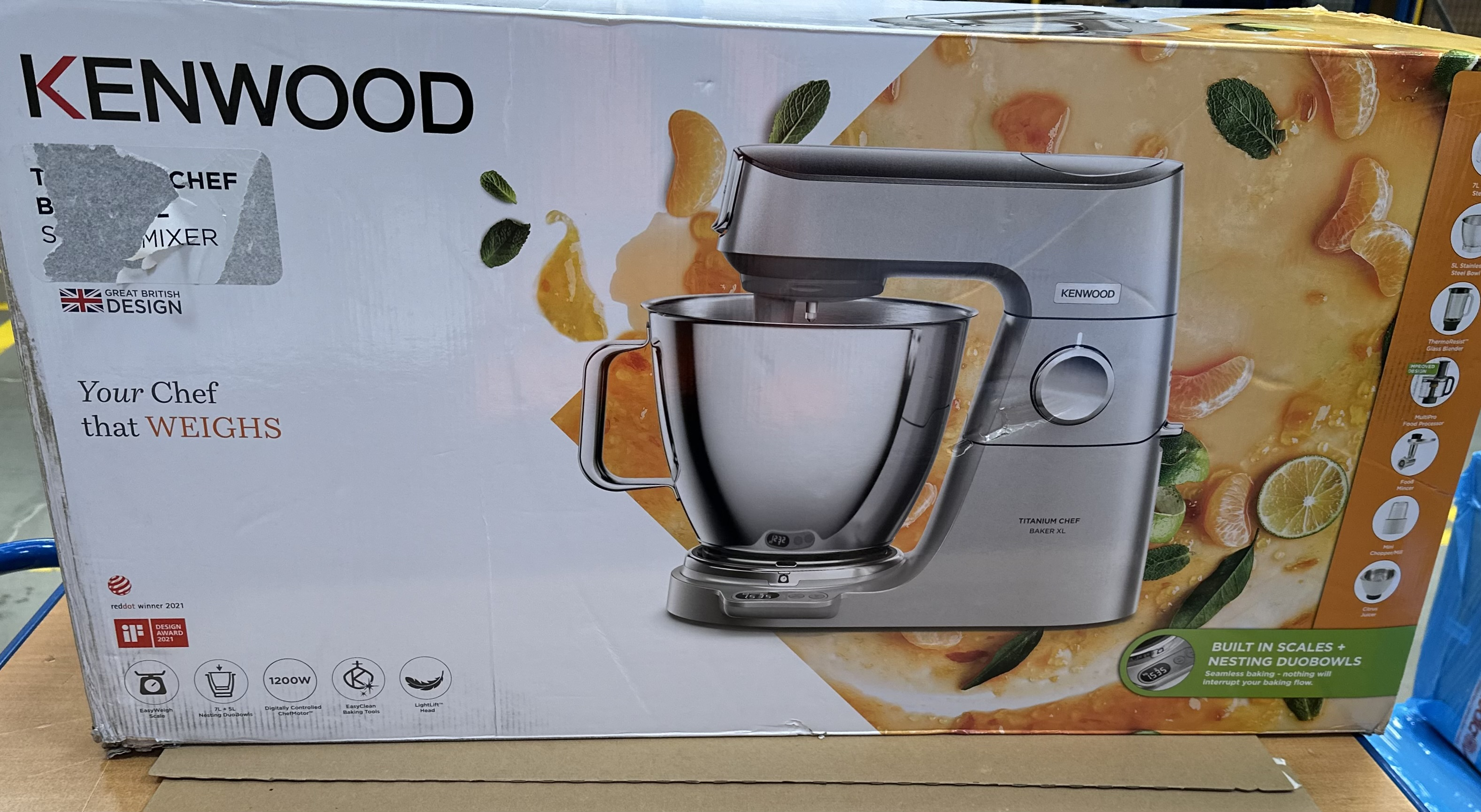 Pudełko Kenwood Titanium Chef Baker XL. Wyświetlany jest srebrny mikser. Pomarańcze i liście w tle.